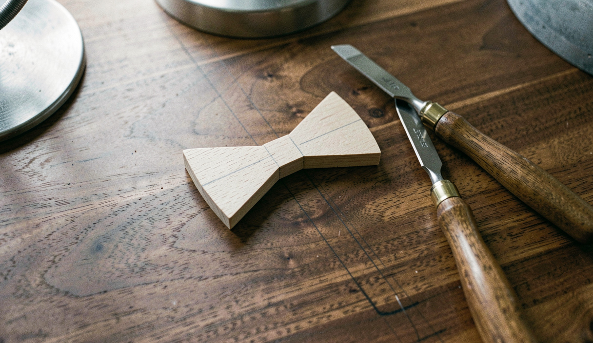 A small bow tie shaped piece of light timber lying on a dark walnut surface with a marking knife and a narrow chisel beside it, pencil layout lines visible, bench lamp light, close view from above.