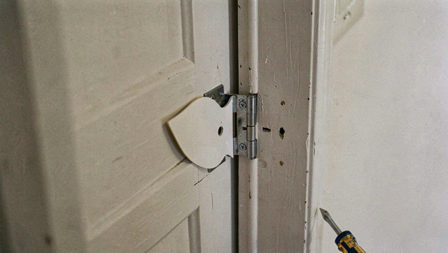 A butt hinge lifted away from its recess in a painted door frame with a piece of card cut to the leaf shape lying in the recess behind it, a screwdriver resting on the frame, close view under hallway light.