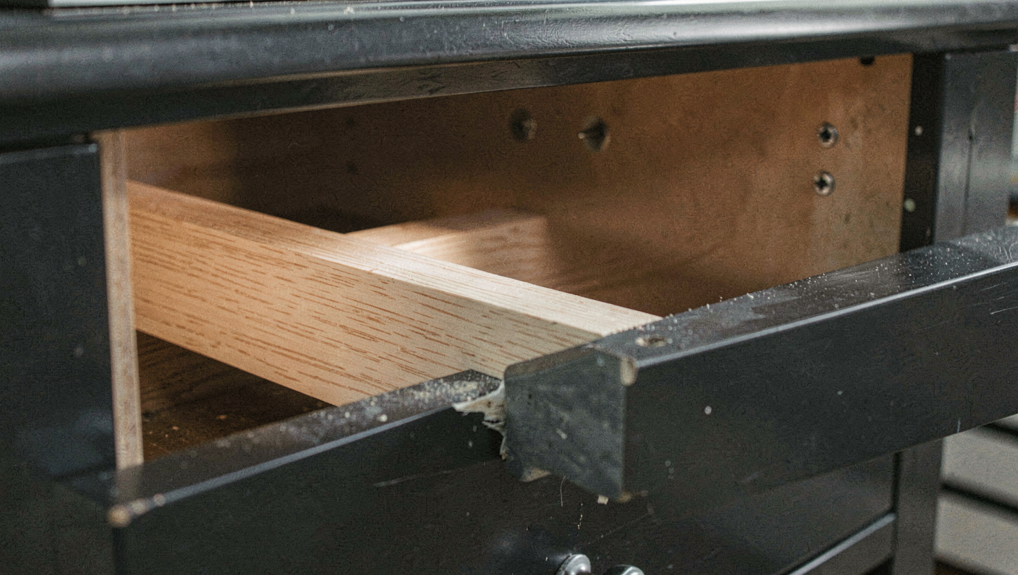 A fresh strip of pale oak seated in a shallow housing inside a dark chest of drawers carcase with a small clamp at the front and two screw heads visible further back, work light shining into the carcase, close view.